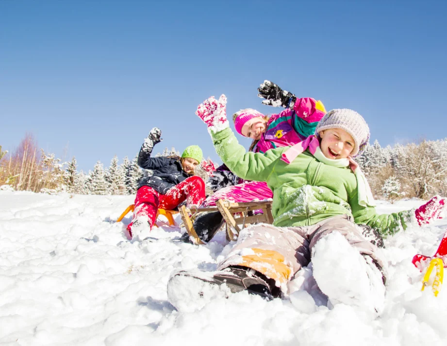 Four children in colorful winter clothing sledding and playing in the snow on a sunny day, with trees and a clear blue sky in the background.