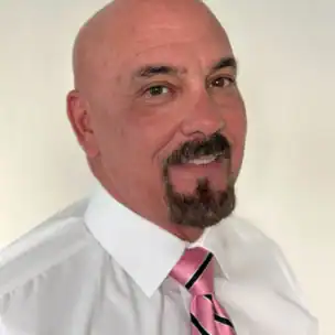 A man with a bald head, goatee, and mustache is wearing a white shirt and a pink striped tie, posing in front of a plain light background.