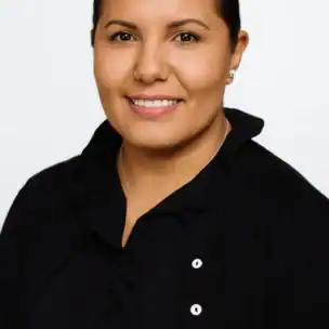 A woman with dark hair tied back, wearing a black buttoned top over a black blouse, poses against a plain white background.