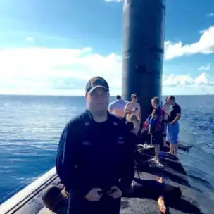 A person in a navy uniform stands on the deck of a surfaced submarine with several people in casual clothing behind him, some near the sail, under a clear sky.