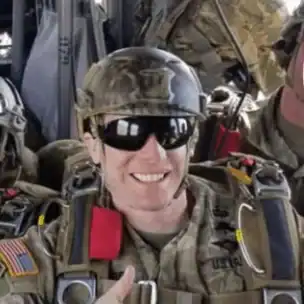A smiling U.S. Army paratrooper in full gear and helmet sits in an aircraft, making a hand gesture and wearing sunglasses.