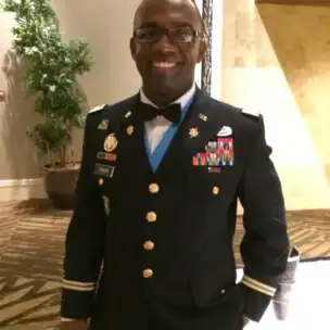 A man in formal U.S. Army dress uniform stands indoors, smiling at the camera. He has short hair, glasses, and is wearing several ribbons and medals on his jacket.