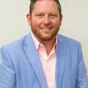 A man with short hair and a trimmed beard, wearing a light blue blazer over a pale pink shirt, stands against a plain light background, smiling at the camera.
