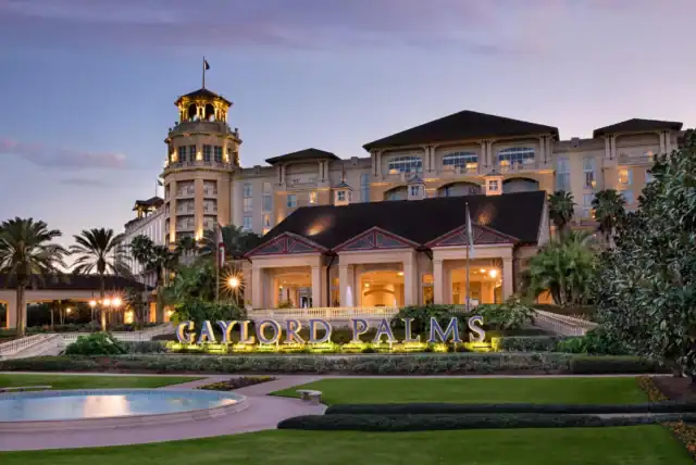 Exterior view of the Gaylord Palms Resort at dusk, featuring a well-lit main entrance, a large hotel building, and landscaped grounds with palm trees.