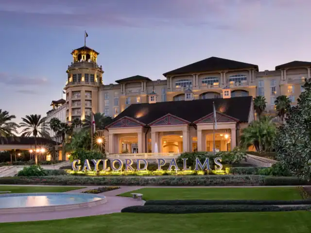 Exterior view of the Gaylord Palms Resort at dusk, featuring a well-lit main entrance, a large hotel building, and landscaped grounds with palm trees.