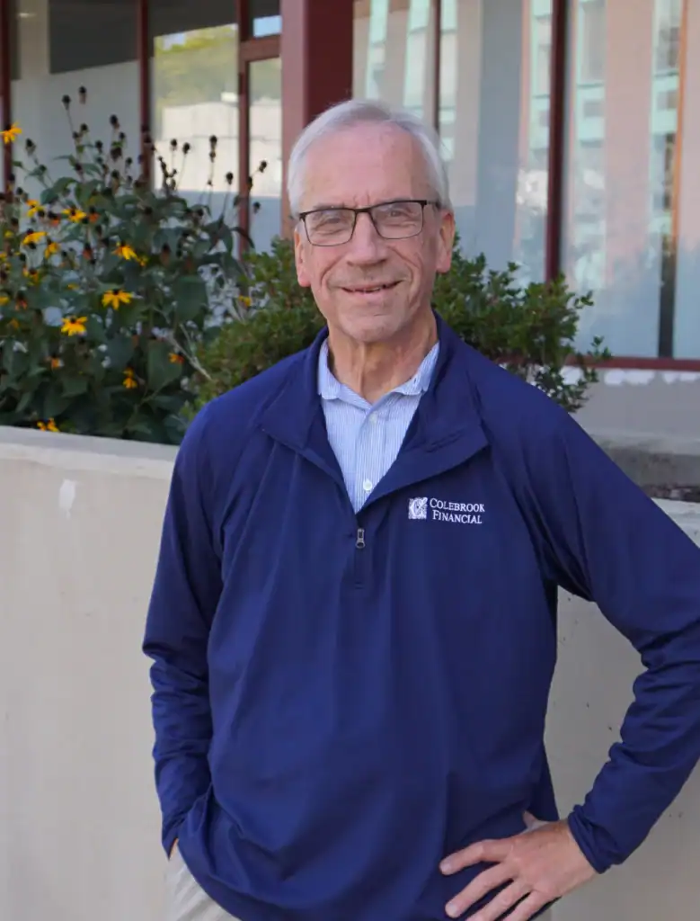 An older man with gray hair and glasses stands outside wearing a blue Courserbook Financial jacket, with plants and a building in the background.