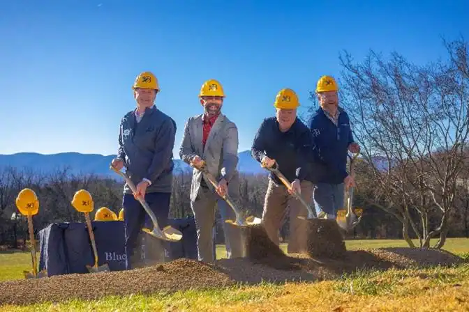 Four people wearing yellow hard hats use shovels to break ground at an outdoor construction site, with trees and mountains in the background.