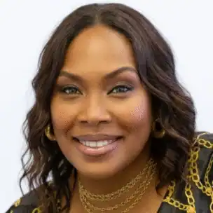 A woman with wavy brown hair, wearing hoop earrings, layered gold necklaces, and a black top with gold patterns, smiling at the camera.