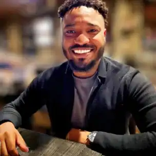 A man with short curly hair and a beard, wearing a dark jacket over a gray shirt, sits at a table and smiles at the camera in a softly lit indoor setting.