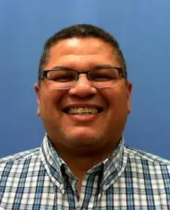 A man wearing glasses and a blue plaid shirt smiles at the camera in front of a plain blue background.