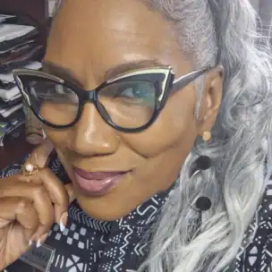 A woman with gray hair in a ponytail, wearing cat-eye glasses, geometric earrings, and a patterned black and white top, smiles at the camera in an office setting.