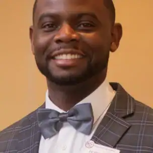 A man wearing a gray plaid suit, white shirt, and gray bowtie smiles at the camera. He has short hair and a trimmed beard, and is wearing a name tag.