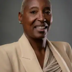 A person with short, light-colored hair wears a beige blazer and draped blouse, smiling in front of a plain gray background.