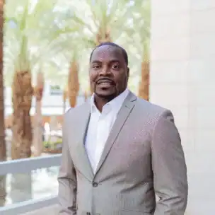 A man in a light gray suit stands outdoors near a stone wall and railing, with palm trees and greenery in the background.
