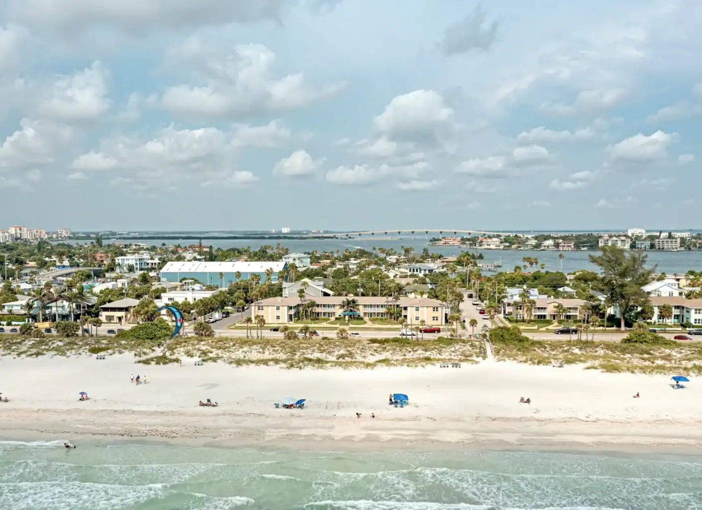 A sandy beach with scattered people and umbrellas, bordered by grass, low-rise buildings, and a body of water with a distant bridge under a partly cloudy sky.