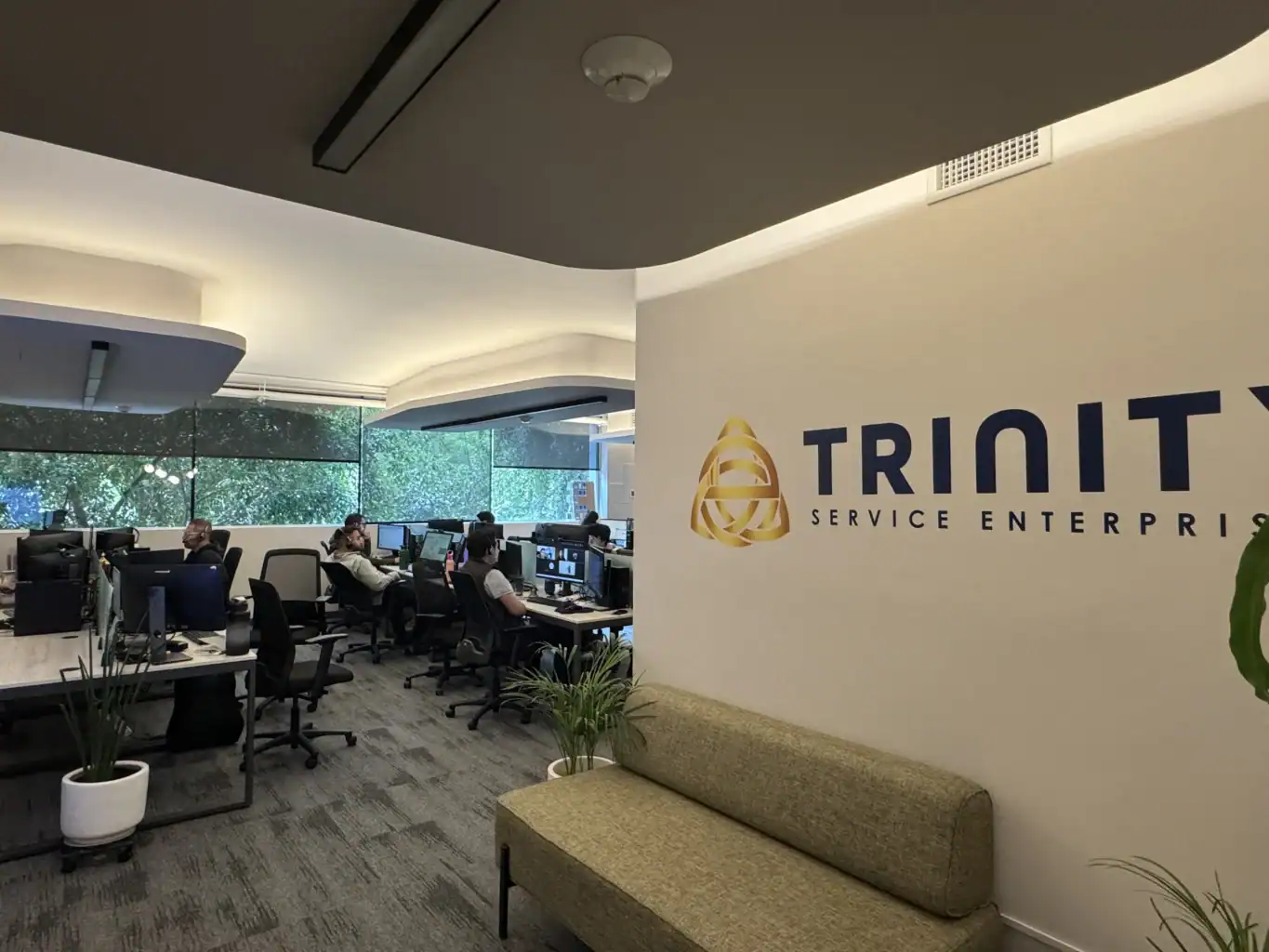 Office with several people working at desks, computers, and monitors; large windows with greenery outside; “TRINITY SERVICE ENTERPRISES” logo on wall; couch and plant in foreground.