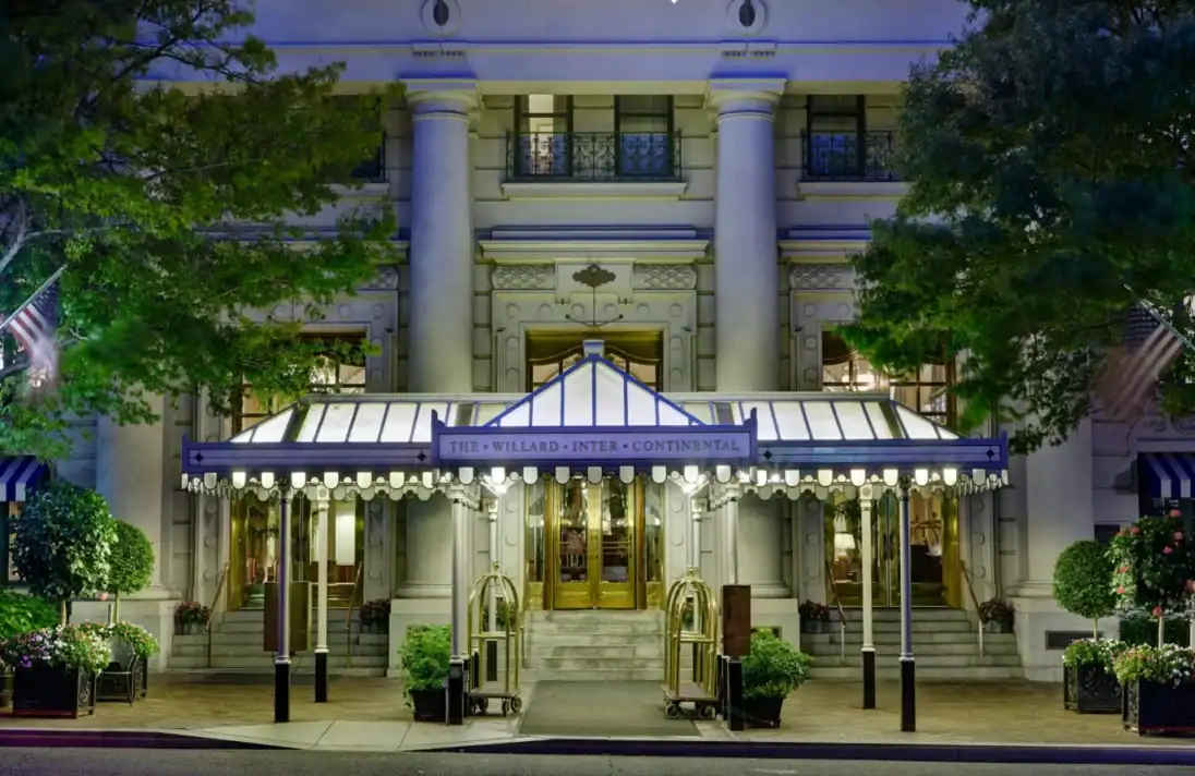 The entrance of the Willard InterContinental Hotel features a lit canopy, revolving doors, potted plants, and ornate columns, seen at dusk.