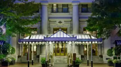 The entrance of the Willard InterContinental Hotel features a lit canopy, revolving doors, potted plants, and ornate columns, seen at dusk.