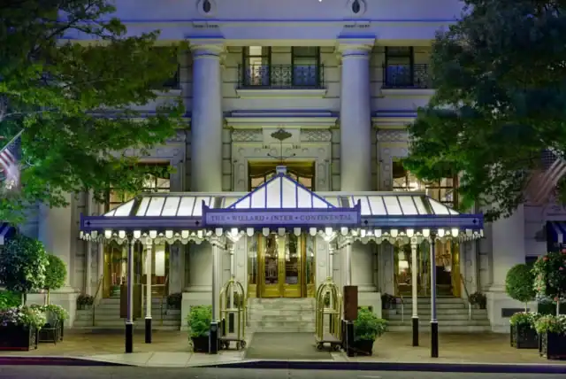 The entrance of the Willard InterContinental Hotel features a lit canopy, revolving doors, potted plants, and ornate columns, seen at dusk.