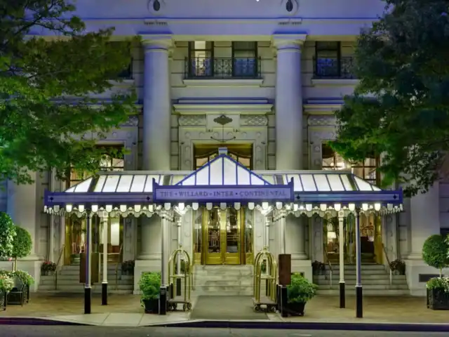 The entrance of the Willard InterContinental Hotel features a lit canopy, revolving doors, potted plants, and ornate columns, seen at dusk.