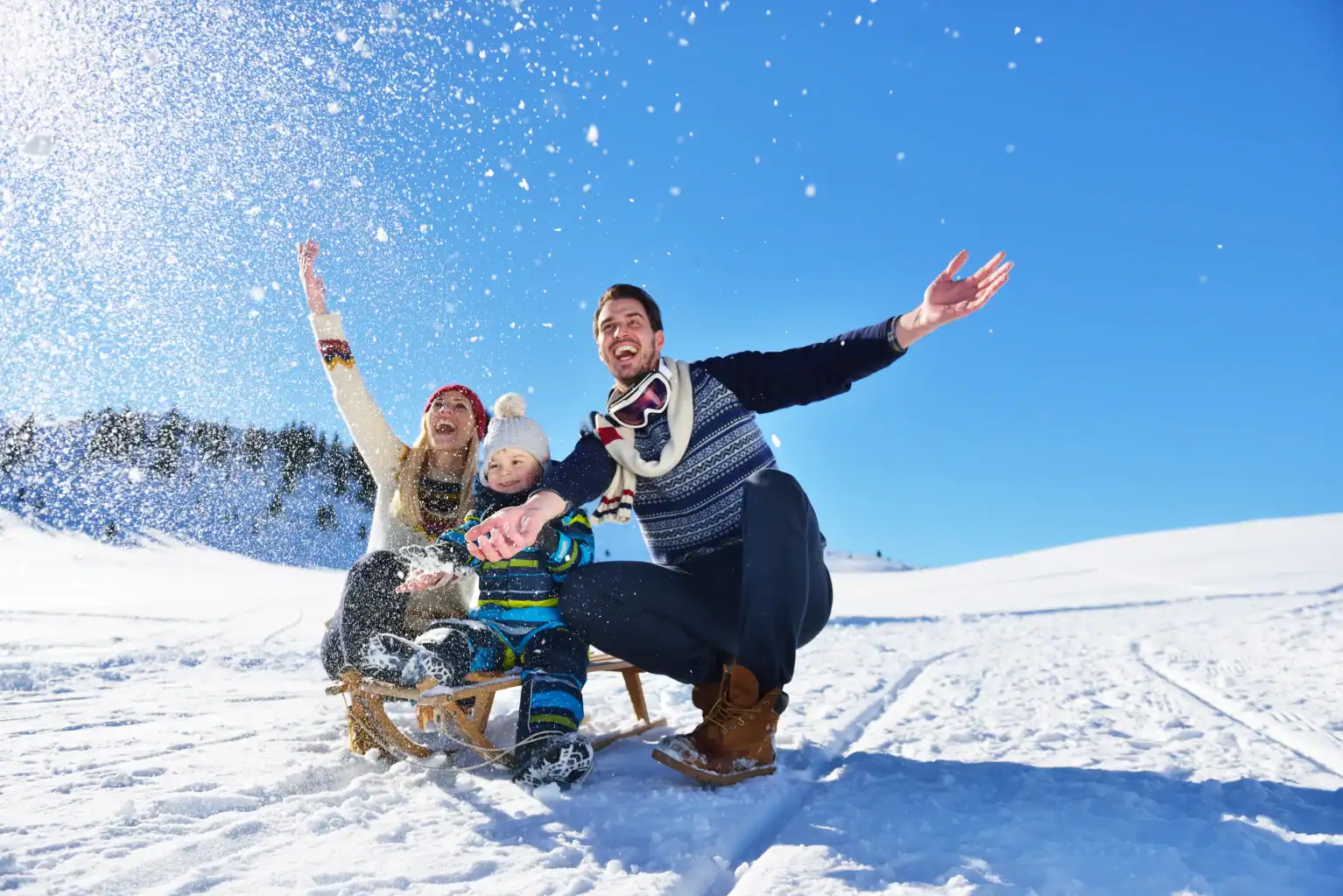 Three people in winter clothing sit on a wooden sled in the snow, arms raised, with snow spraying around them under a clear blue sky.