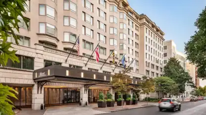 Hotel entrance with multiple flags above, large windows, and potted plants along the sidewalk; a car drives past on the street in front.
