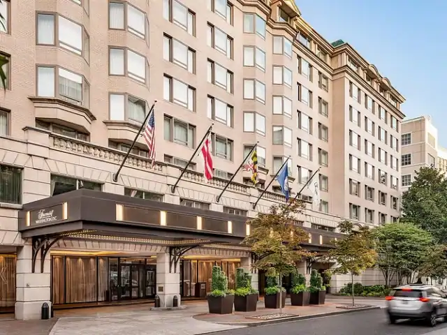 Hotel entrance with multiple flags above, large windows, and potted plants along the sidewalk; a car drives past on the street in front.